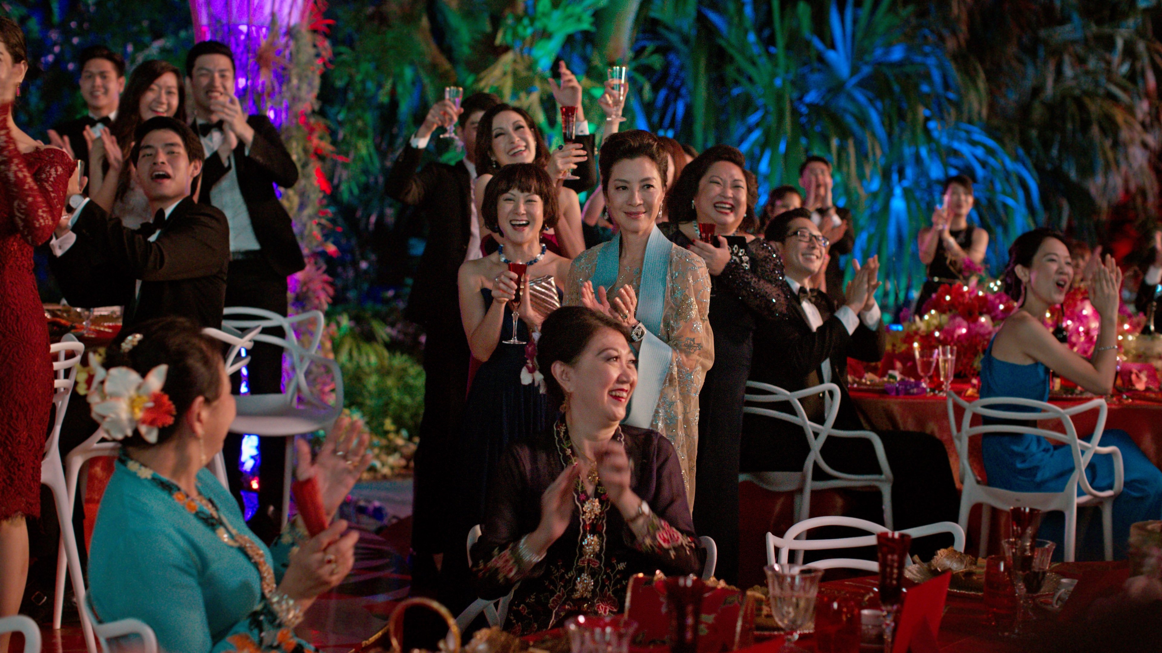 A wide ensemble frame suggesting a grand family celebration, dozens of figures in traditional embroidered clothing beneath a chandelier in a marble hallway, one woman in the foreground laughing mid-conversation, warm amber light, opulent production design